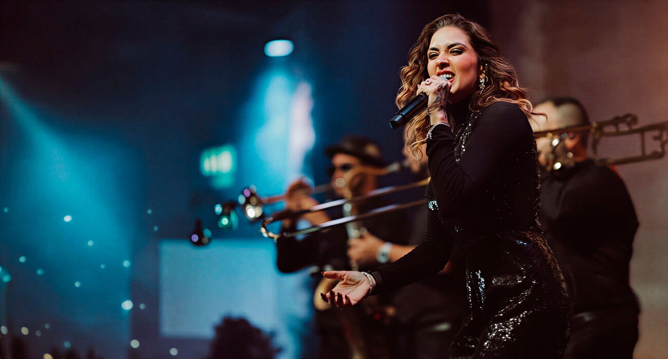 Vocalista da Banda Prime cantando com intensidade ao lado do naipe de metais no palco
